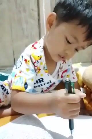 A young child with dark hair, wearing a white t-shirt with a colorful pattern featuring cartoon airplanes and stars, is sitting on a surface that appears to be a bed with a wooden plank background. The child is holding a green pen in their right hand and is in the process of writing or drawing on a notebook that is open in front of them. The child's gaze is directed downwards towards the notebook. To the right of the child, a tan-colored stuffed animal is visible. The lighting suggests an indoor environment.