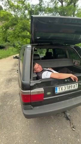A person lies in the open trunk of a black Range Rover parked on a dirt road. The person is wearing a white t-shirt and a dark vest. The trunk is open, and the rear of the vehicle is facing the camera. The license plate reads TX 3189 AT. Trees and a rocky incline are visible in the background.