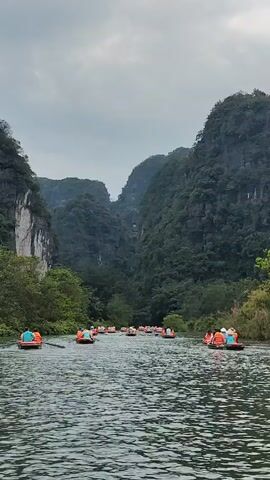 A flotilla of boats carrying tourists wearing orange life vests are traveling on a river flanked by steep, forested mountains. The sky is overcast, casting a somewhat dull light on the scene. The water surface has a moderate ripple, reflecting the gray sky. The boats are positioned in the center of the river, moving towards a narrow opening between two large rock formations. The surrounding land is densely vegetated with trees and bushes.