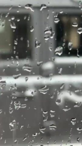 The video shows a view through a rain-covered window. Initially, the focus is on the raindrops on the glass, blurring the background. As the camera adjusts, a silver or grey Land Rover Defender comes into focus parked on the street outside. The vehicle is partially covered in snow or slush, suggesting wintry conditions. Buildings are visible in the background, one of which appears to house a bar. The overall scene conveys a cold and wet day.
