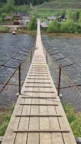 The camera is positioned at the beginning of a long suspension bridge, looking directly down its length. The bridge is constructed of wooden planks running lengthwise, with smaller planks laid across to create a tread. Metal cables and netting form the sides of the bridge. The river below is visible, with dark water and small ripples. On the opposite bank, there are several houses and buildings, surrounded by green trees and grass. Further in the background, a forested hill rises. There is no one visible on the bridge.