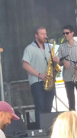 A man with a beard, wearing a light blue collared shirt and dark blue jeans, stands on a stage playing a tenor saxophone. He is facing slightly to the left of the camera and looking down at the saxophone. To his right, another person wearing sunglasses and a patterned shirt holds a trumpet and appears to be looking at the saxophonist. They are both on a stage with a dark backdrop and visible lighting equipment. In the foreground, the heads of audience members are visible, with one person wearing a red hat. The overall environment appears to be an outdoor performance.