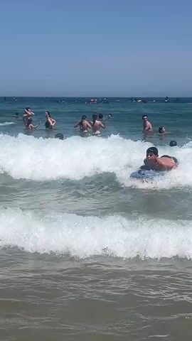 A group of people are at the beach on a sunny day. Several individuals are in the water, some of them swimming or playing in the waves. One person, a young male, is on a boogie board in the foreground, riding a wave. Another young male in the background is also on a boogie board. There are more people in the water further out, and a rocky structure is visible in the distance. The water is blue and clear, with white foam from the waves. The sky is bright and clear.