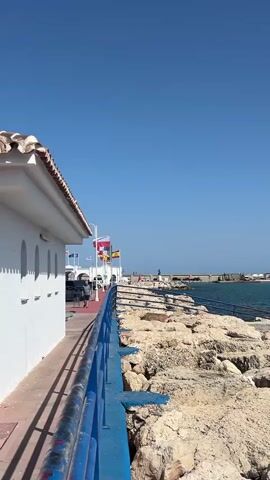 A white building with a tiled roof runs along the left side of the frame. A blue railing runs along the right side of the building, separating it from a rocky embankment that slopes down to the sea. Several flags are visible beyond the railing, and in the distance, a breakwater extends into the sea, with a lighthouse at its end. The sky is clear and blue.