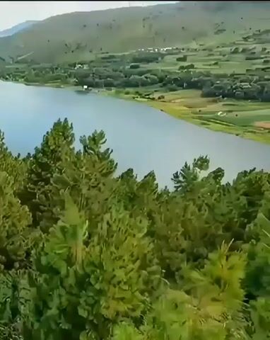 The video shows a panoramic view of a natural landscape with a large lake in the center. In the foreground, there is a dense forest of green trees. Beyond the lake, the land is characterized by rolling hills covered in lush vegetation. In the distance, a mountain range is visible under a partly cloudy sky. The scene appears tranquil and expansive, showcasing a serene natural environment. There are no people or specific objects that suggest human activity in this particular frame.