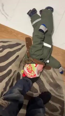 Here is a detailed description of the scene in the video: 

The video appears to be taken from a first-person perspective, looking down at a small child on the floor. The child, wearing a green winter snowsuit, colorful hat, mittens and shoes, is lying on their back on a round zebra-striped rug. Their head is near the edge of the rug, resting on the boot of a person wearing jeans and black shoes. The person is standing next to the rug. Part of the floor appears to be white tiles and the rest is wood.