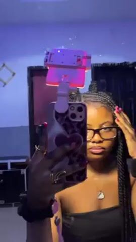 A person with dark skin, braids, and glasses is standing in a room and holding up their phone to take a selfie in a mirror. The phone has a pink case with a heart design and an attached pink light fixture. The room has dim, colorful lighting and various objects scattered around, including speakers, a framed decoration, and a door. The person makes a hand gesture towards the camera.
