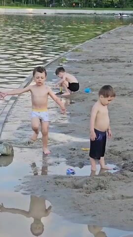This video depicts three young boys at a beach or lakeside. The setting is a sandy shore with shallow water. In the foreground, a boy wearing white patterned swim trunks walks through the shallow water, splashing his feet. To his right, another boy in dark blue swim trunks stands on the wet sand, looking down. In the background, a third boy is crouched on the sand, playing with toys, including a yellow shovel and a blue bucket. The water appears calm, with gentle ripples. The lighting suggests it is daytime, possibly late afternoon or early evening, given the soft light. The overall atmosphere seems relaxed and playful, with the children enjoying their time outdoors. There is no visible text in the video. The location context provided is Nizhnyaya Saniba, Russia.