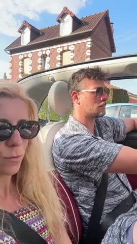 The video shows a woman and a man in a convertible car. The woman, in the foreground on the left, has long blonde hair and is wearing sunglasses. The man, in the passenger seat on the right, is wearing sunglasses and a grey patterned shirt. They appear to be driving down a street with houses and trees on either side. The sky is blue and clear. The car is red on the interior. The man's right arm is on the car door.