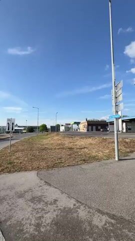 The video pans across a dry, grassy area next to a paved walkway. In the foreground, a road sign with a bicycle symbol and arrows indicating lanes is visible. Behind it, a blue-roofed structure, possibly a gas station or car wash, is in the distance with several bays. Other buildings, including a brown one with a mural, and streetlights, are also present. The sky is clear blue with scattered white clouds. The ground is covered in dry grass and some weeds. There are no people visible in the video. The camera is in motion, suggesting it is being held by someone walking or riding.