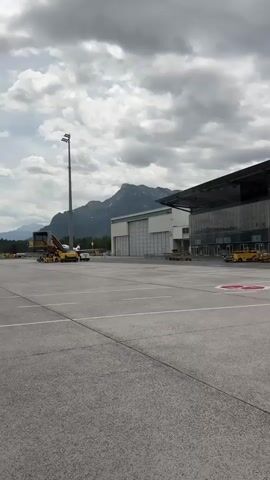 The media file captures an airport tarmac scene with a modern terminal building and hangars in the background. Mountains loom in the distance under a cloudy sky, suggesting an overcast day.

On the tarmac, several yellow airport vehicles are visible, including what appears to be a baggage tug and a specialized aircraft handling vehicle. One of these vehicles is positioned near a hangar.

A prominent subject in the foreground is a police officer in uniform, wearing a tactical vest and a hat, and holding a rifle. The word "POLIZEI" (German for "Police") is visible on the vest. Further to the right, a police van, also marked with "POLIZEI" and featuring Austrian license plates, is parked.

The terminal building has a distinctive modern design with a large, sloping roof and extensive glass facade. Multiple doors are visible along the ground floor of the terminal. A circular red marker is painted on the tarmac.

The overall impression is of an operational airport with a visible security presence. The weather appears to be typical for a cloudy day in an alpine region.