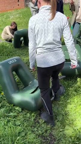 Several people are outside on a grassy field, interacting with green inflatable river tubes. Most of the adults are using a foot to push down on the tubes to inflate them. One adult is seated on the ground working on their tube. The day is sunny, with blue skies and puffy white clouds, and a few buildings are visible in the background.