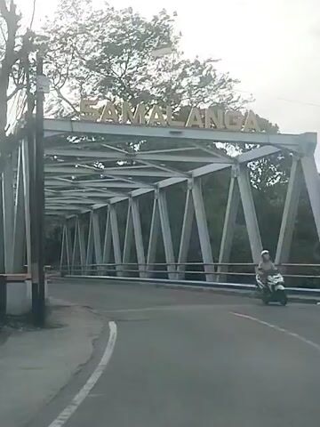 A video captures a vehicle's perspective driving onto a bridge. The bridge is constructed of steel trusses and has the word "SAMALANGA" visible in large letters above its entrance. On the bridge, a large white truck with a red cab is proceeding in the same direction as the camera vehicle. Trees and foliage are visible on either side of the road beyond the bridge.