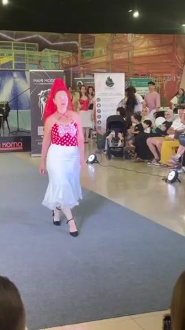 A woman is walking down a runway wearing a red and white polka dot halter top, a white skirt with lace trim, and a red veil. She is performing a dance with hand movements. Spectators are seated on the right side of the runway, and there are banners and signs in the background. The environment appears to be an indoor mall or event space.