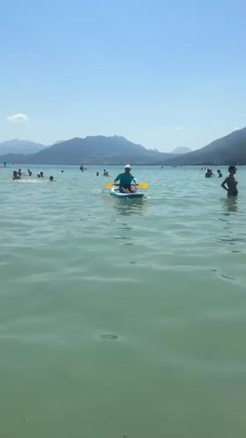 A person on a paddleboard is paddling on a lake. The person is wearing a blue long-sleeved shirt, brown shorts, and a white baseball cap. They are kneeling on the paddleboard and using a yellow paddle. There are other people swimming and playing in the water in the background. Mountains are visible in the distance under a clear blue sky. The water is a light greenish-blue color and appears calm. The sun is shining brightly.