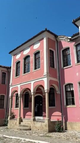 The video shows a street scene with two pink buildings on a sunny day. The buildings have white trim and arched doorways. The street is paved with cobblestones. Trees and a blue sky are visible. There are no people in the video.