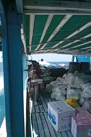 THE VIDEO SHOWS THE INTERIOR OF A BOAT, LIKELY A CARGO VESSEL, LOADED WITH NUMEROUS SACKS AND BOXES. A MOTORCYCLE IS PARKED ON THE LEFT SIDE OF THE BOAT. THE BOAT APPEARS TO BE IN MOTION, AS EVIDENCED BY THE MOVEMENT OF THE WATER VISIBLE TO THE LEFT. THE CEILING OF THE BOAT IS COVERED WITH GREEN FABRIC STRETCHED OVER A FRAMEWORK OF WOODEN PLANKS. THE FLOOR OF THE BOAT IS MADE OF WOODEN PLANKS. THE CARGO INCLUDES SEVERAL LARGE, LIGHT-COLORED SACKS, SOME BLACK DRUMS, AND VARIOUS BOXES, INCLUDING ONE WITH THE TEXT "COLD STORAGE" AND ANOTHER WITH WRAPPING THAT APPEARS TO BE PACKAGES OF MONEY. THE OVERALL IMPRESSION IS A DENSELY PACKED CARGO HOLD ON A VESSEL TRANSPORTING GOODS ACROSS THE WATER.