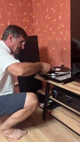 A man with glasses wearing a white shirt and blue shorts is kneeling on a wooden floor. He is reaching out to a turntable, where he is holding a record. The turntable is on a wooden shelf with black legs. A black speaker is to the left of the turntable, and a black electronic device is below it. The wall behind the man has a red floral wallpaper. The scene is lit by artificial light.