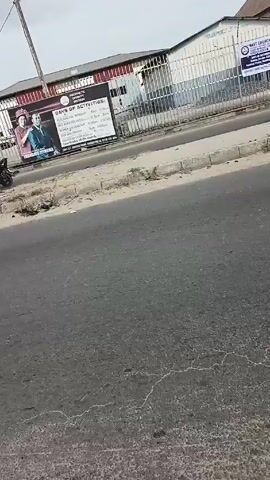 The video shows a street scene in what appears to be a developing country. The street is asphalt, patched and faded, with a sandy shoulder. The sky is overcast. Vehicles, including cars, auto rickshaws (tuk-tuks), and motorcycles, are seen moving along the road. The sidewalks are partially paved, and there are buildings and fences along the sides of the road. Power lines and poles are visible overhead. A large advertisement for a religious event is displayed on a fence. The overall impression is of a busy, somewhat chaotic, urban environment.