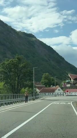A person wearing a light-colored t-shirt and dark pants walks across a bridge. In the background, there are several buildings, a large mountain covered in trees, and a sign that reads "I Love Savnik." Two motorcycles are parked near the sign. The sky is partly cloudy.