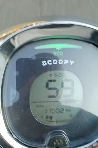 The video begins with a close-up of a motorcycle's digital dashboard, displaying "SCOOPY" and an "ECO" indicator. The speed reads 59, and the odometer shows 11002. The camera then pans out to show the road ahead, with a white car visible in the distance. The motorcycle appears to be moving at a steady pace on a paved road lined with trees and a guardrail. Another motorcycle with a rider is ahead, and the video continues to show the perspective from the moving motorcycle as it travels down the road, with the dashboard still visible.