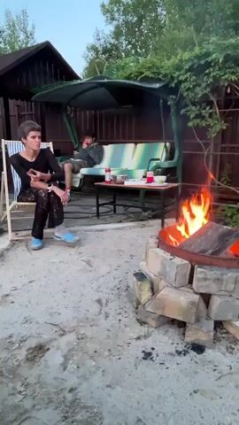 The video opens with a view of a backyard patio area. In the foreground, a fire burns in a circular fire pit constructed from bricks. To the left, a person sits in a striped deck chair, looking to the right. Further back, a green swing set with a striped cushion is visible. On the swing set, a second person is seated, facing towards the fire pit. A small table is positioned in front of the swing set, holding various items, including cups and containers. The background consists of a wooden fence and trees. The overall scene depicts an outdoor gathering with a fire.