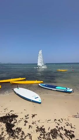 This media file captures a serene beach scene in Castiadas, Italy. The foreground is dominated by a sandy beach scattered with dried seaweed and debris. Several watercraft are visible, including a white and blue stand-up paddleboard resting on the sand, another paddleboard with a blue and green design partially submerged in the water, and three bright yellow kayaks lined up near the shore.

In the water, a small white sailboat with its sails up is gliding across the calm, clear blue sea. Another yellow floating object, possibly a buoy or another small inflatable, is visible further out. The sky is a clear, vibrant blue, indicating a sunny day with pleasant weather. The sun appears to be high in the sky, suggesting it is daytime, likely late morning or early afternoon. The overall atmosphere is peaceful and inviting, evoking a sense of tranquility and the enjoyment of water activities. There is no visible text in the media file.
