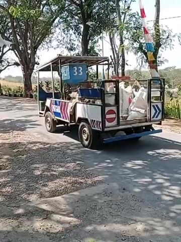 A small, open-air vehicle with a white body and a blue frame, marked with the number 33 on a blue panel, drives along a paved road. The vehicle has a roof supported by poles, and a cargo bed filled with several large, white bags. A person is seated in the driver's seat. On the sides of the vehicle are signs, including a circular red sign with a white horizontal bar, indicating "no entry" or a prohibition. There are also blue and white striped markings along the sides. Flags, one red and white and another with red, white, and blue stripes, are visible, as well as a sign with a blue arrow pointing to the right. The vehicle is surrounded by trees, with lush green foliage in the background. The road appears to be bordered by gravel and some patches of grass. The scene is outdoors and appears to be during the daytime, with sunlight casting shadows.