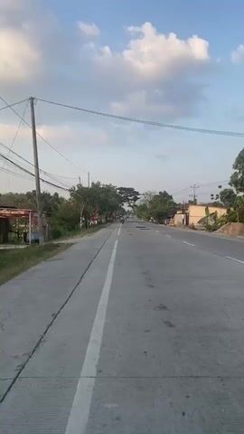 The video shows a paved road with two lanes and a white line dividing them. The road is lined with trees and some buildings on either side. The sky is blue with some white clouds. There are a few people on motorcycles on the road in the distance. The overall scene appears to be a rural or suburban area. The video is shot from a moving vehicle.