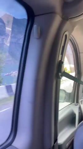 The video shows a view from inside a vehicle, looking out the window at a scenic mountain landscape. In the foreground, a road is visible with concrete barriers along the side. Beyond the road, a large body of turquoise water, likely a lake or reservoir, stretches towards imposing mountains. The mountains are steep and rocky, with some sparse vegetation on their slopes. The sky is overcast with dramatic clouds, suggesting either early morning or late evening. There are some structures on the far side of the water, including what appears to be a small settlement or resort area with buildings and boats. The camera is moving, indicating the vehicle is in transit. The overall impression is one of natural beauty and a journey through a mountainous region.