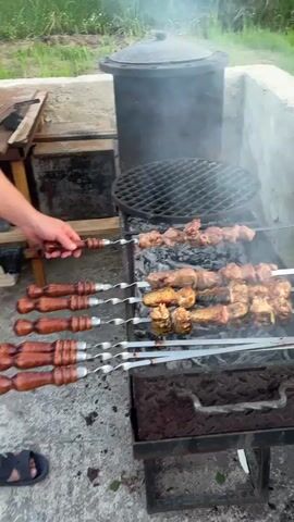 A close up shot shows a grill station, seemingly outdoors with grass and shrubbery in the background. A person wearing blue sandals stands beside the grill. The grill itself is a DIY concrete structure with a metal grill surface covered with hot coals. On the grill are several skewers, some holding pieces of meat and some holding what appears to be cooked fish. Smoke rises from the food and the coals. There is a large metal pot placed towards the back of the grill area.