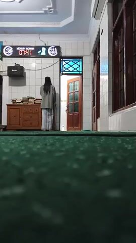 A person is standing in a room with a green carpet. The person is facing a wall with a digital clock that shows the time 07:36. The person is wearing a grey jacket, a white sarong, and a black prayer cap. There is a wooden cabinet to the left of the person, with books stacked on it. There is a door to the right of the person, which is slightly ajar. The room appears to be a prayer room or a mosque.