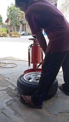 A person is using a tire changing machine to remove a tire from a rim. The person is wearing a maroon t-shirt and black pants with blue stripes. They are also wearing sandals. The tire is black with a silver rim. The tire changing machine is red. The scene appears to be outdoors on a paved surface. There is a house and trees in the background.