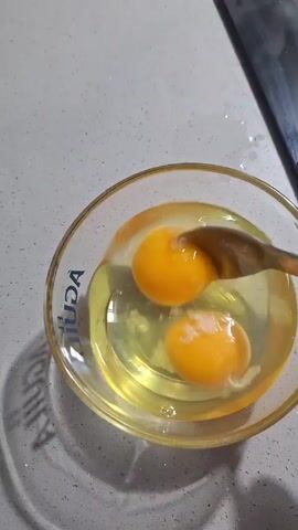 The video shows a small clear glass bowl containing three raw eggs on a gray countertop. A metal spoon is used to vigorously whisk the eggs, blending the yolks and whites together. The bowl has a gold rim and the word "AGUILA" printed on the side. The video is shot from directly above, providing a clear view of the mixing process. A small portion of a black object is visible in the top right corner of the frame.