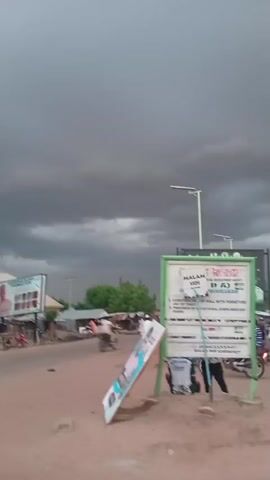 A man wearing a pink shirt and a dark baseball cap walks towards the camera on a dirt road. In the background, there are trees, buildings, and several motorcycles. The sky is dark and cloudy, suggesting an impending storm. The camera is shaking and the image is blurred.