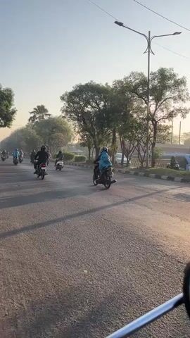 The video shows a street scene with many motorcycles on a paved road. The motorcycles are moving in the same direction. Most of the riders are wearing helmets. The sun is low in the sky, casting long shadows. There are trees and streetlights on the side of the road. The overall atmosphere is that of a busy commute.