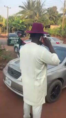 The video depicts a man in a white shirt and a maroon hat talking on a phone next to a silver car. The camera pans to show a group of people walking on a dirt road, some dressed in traditional attire. Several cars are parked nearby, and a motorcycle drives by. The environment appears to be a residential area with trees and foliage.