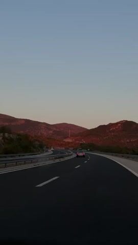 A car's perspective of a highway at dusk. The road curves to the left, with guardrails on either side. In the distance, mountains are visible, illuminated by the setting sun, casting a red glow. A few other vehicles, including a truck and a car, are also on the highway. The sky is a clear, pale blue.