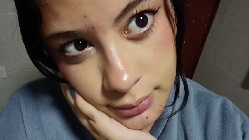 A close-up video shows a young woman with dark hair and brown eyes. She is wearing a light blue top. Her face is lightly made up, with noticeable eyeliner and a touch of blush. In the first shot (0:00-0:01), she rests her chin on her hand, looking slightly to the side with a subtle smirk. Her fingernails are long, pointed, and painted a sparkling teal color. In the second shot (0:01-0:04), she brings her hand up to her cheek, touching it gently with her teal-colored nails, and smiles more openly, looking towards the camera. The background consists of light-colored tiled walls, suggesting an indoor setting. There is no visible text.