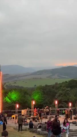 This is a vertical, full-shot video of an outdoor music festival at dusk. In the foreground, a crowd of people sits and stands on tiered seating, facing a stage in the middle ground. The stage has several musicians, musical instruments, and lighting rigs. Behind the stage, a lush green forest stretches across rolling hills, and in the background, distant mountains are visible under a cloudy, orange-tinted sky. The lighting from the stage and sky casts a warm glow on the scene.