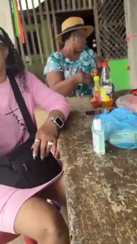 Two women are seated at a weathered, wooden table in an outdoor setting. The woman in the foreground is wearing a light pink dress, glasses, and a black fanny pack. She has several rings on her fingers and an Apple watch on her left wrist. She is holding a reddish, exotic-looking fruit in her right hand and appears to be eating it.  The other woman, seated further back, wears a patterned blouse and a straw hat. She is also holding something in her hand, possibly food. The table has bottles of juice, a mobile phone, and a bottle of hand sanitizer on it.  The background features what seems to be a rustic structure made of wood. There are red, green, and yellow decorative streamers hanging from the structure.