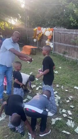 In an outdoor setting on a sunny day, a man is seen throwing money on a lawn where several young boys are gathered. The children, dressed in various casual clothes, are scrambling to pick up the bills. One boy is wearing a medal around his neck. In the background, there's a balloon arch, a table, and other people, suggesting a party or celebration. A wooden fence marks the boundary of the yard. The lighting indicates it may be late afternoon.