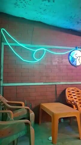 The video pans across a brick wall with a neon green light strung in a wavy pattern. To the right, a circular sign with a blue and white design is mounted. In the foreground, there are several orange plastic chairs and a small orange plastic table. The floor is concrete. In the background, through an opening, a dimly lit area with more chairs, tables, and people can be seen. Motorcycles are also visible in the background. The lighting is artificial and appears to be from the neon light and ambient lighting in the background.