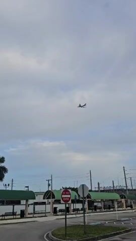 An airplane flies across a cloudy sky. Below, there are palm trees, covered waiting areas with benches, and a street. A red and white "DO NOT ENTER" sign is visible in the foreground.