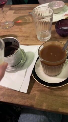 A hand pours a light-colored liquid from a small clear glass into a clear glass mug filled with dark coffee. The mug is on a white saucer with a black and gold rim. A small silver spoon is in the mug. A white napkin and a metal coffee filter are next to the mug. A patterned plate with an empty small bowl, a plain glass, and a small purple bowl are in the background. The surface is a light-colored wooden table. The person is wearing blue jeans.