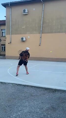 The video shows a man on a concrete basketball court hitting tennis balls. He is wearing a short-sleeved shirt with a lion design, black shorts, a white baseball cap, and orange running shoes. He is hitting the tennis balls with a black tennis racket towards the back of the court. The basketball court is next to a light tan colored building with two air conditioning units mounted on the wall.  A basketball hoop with a torn net is visible on the right side of the building. There is gravel around the basketball court. The sky is visible at the top of the frame.