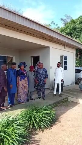 The video opens with a view of a building with a white exterior and a brown-tiled roof. In front of the building, five people are gathered on a concrete porch. From left to right, the individuals are a man in a blue outfit and a blue cap, a woman in a patterned blue and white dress with a matching headwrap, a man in a gray outfit and a red cap, a man in a patterned blue and white outfit, and a man in a white outfit. A car, a silver minivan, is parked to the right of the building, and a light blue minivan is parked further to the right. The ground in front of the building is a mixture of dirt and gravel. The video then pans to the right, showing the side of the building and the parked minivans. A man in a checkered shirt and dark pants is standing near the light blue minivan, holding a camera or phone. The video continues to pan, showing more of the minivans and the surrounding trees. Finally, the video pans back to the left, returning to the group of people standing on the porch.
