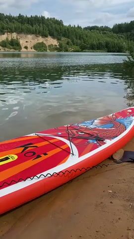 A paddleboard with a red and white koi fish design rests on a sandy shore next to a lake. A person wearing black swim trunks stands in the water with their back to the camera. The lake is surrounded by green trees and a sandy cliff face under a cloudy sky.