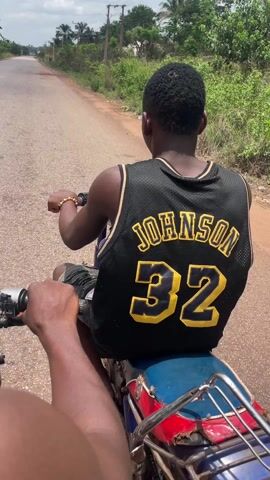 Two people are riding on a motorcycle down a paved road. The person in front, who is driving, is wearing a black and yellow tank top and a black baseball cap. The person in the back is wearing a black and yellow basketball jersey with the name "JOHNSON" and the number "32" on the back. The motorcycle is old and has a metal rack on the back. The road is surrounded by trees and vegetation. The sky is partly cloudy.