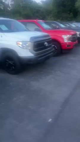 The video shows a line of trucks parked on asphalt. The first truck is white and appears to be a Toyota Tundra. The second truck is red and looks like a Ford F-150. Several other trucks are visible behind these two, all parked in a row. The lighting suggests it is either early morning or late evening. There are trees and foliage in the background. The camera is moving slightly, showing a close-up view of the front of the trucks. No people are visible.